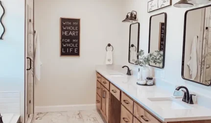 A modern bathroom features a double vanity with two mirrors, a shower, and a framed sign on a light wall.
