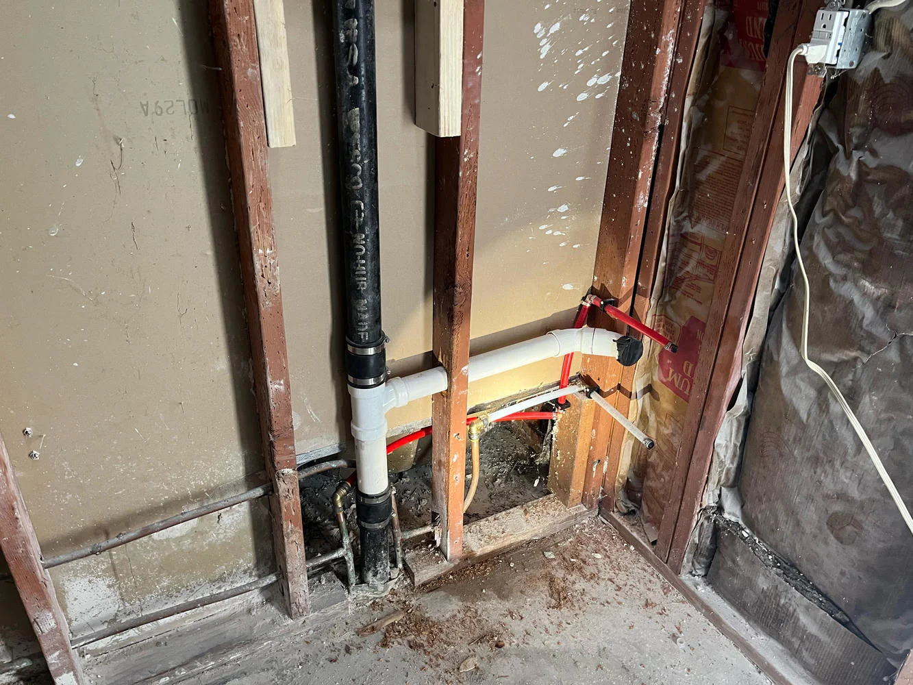 Plumbing pipes exposed in a wall with insulation, showing a black drain pipe, white PVC, and red PEX.